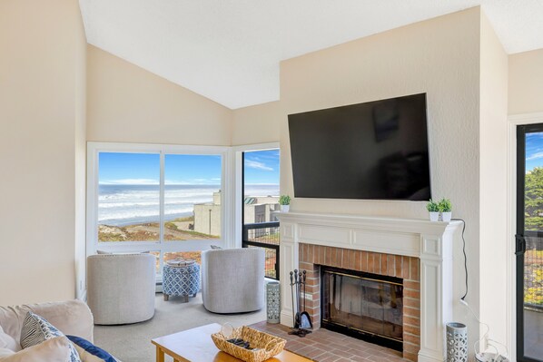 A living room with a fireplace, a TV, and a large window overlooking the ocean. - Beach Condo Pelican 14 (Pajaro Dunes)