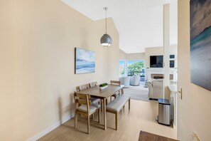 A modern dining area with a wooden table and chairs, a hanging pendant light, and a painting on the wall. - Beach Condo Pelican 14 (Pajaro Dunes)