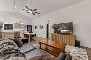 A modern living room with a sofa, coffee table, TV, and kitchen in the background. - Cool Pines Retreat | AC + Large Porch| Sleeps 6 (Pinetop Country Club)