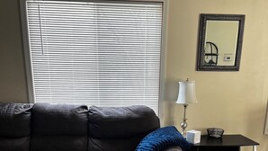 Un salon avec un canapé en cuir foncé, un pouf à motifs, un tapis noir et blanc, une petite table avec une lampe et une fenêtre avec des stores.