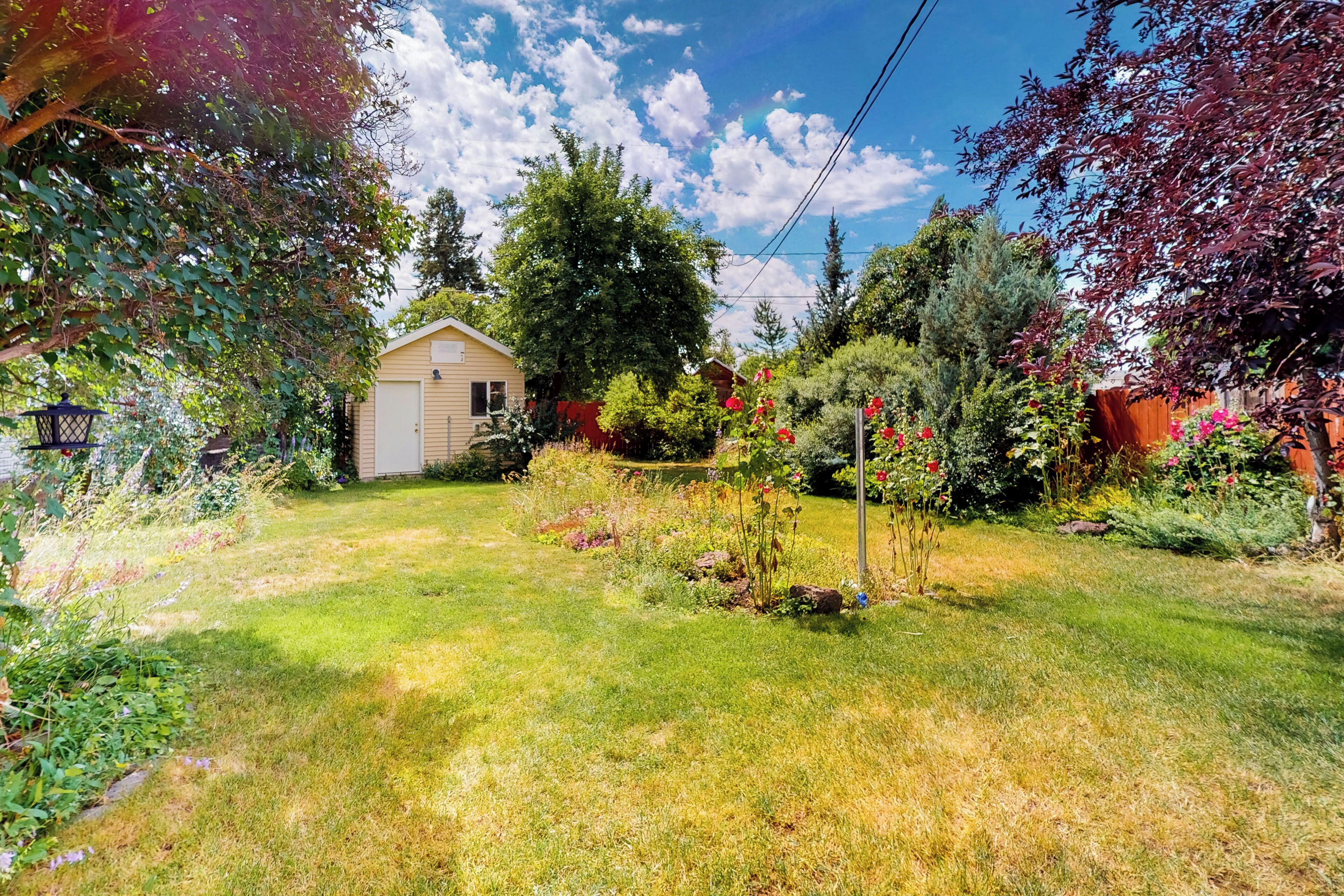 Un jardin bien entretenu, avec une grande variété de plantes et de fleurs, un petit abri de jardin et un ciel bleu dégagé.