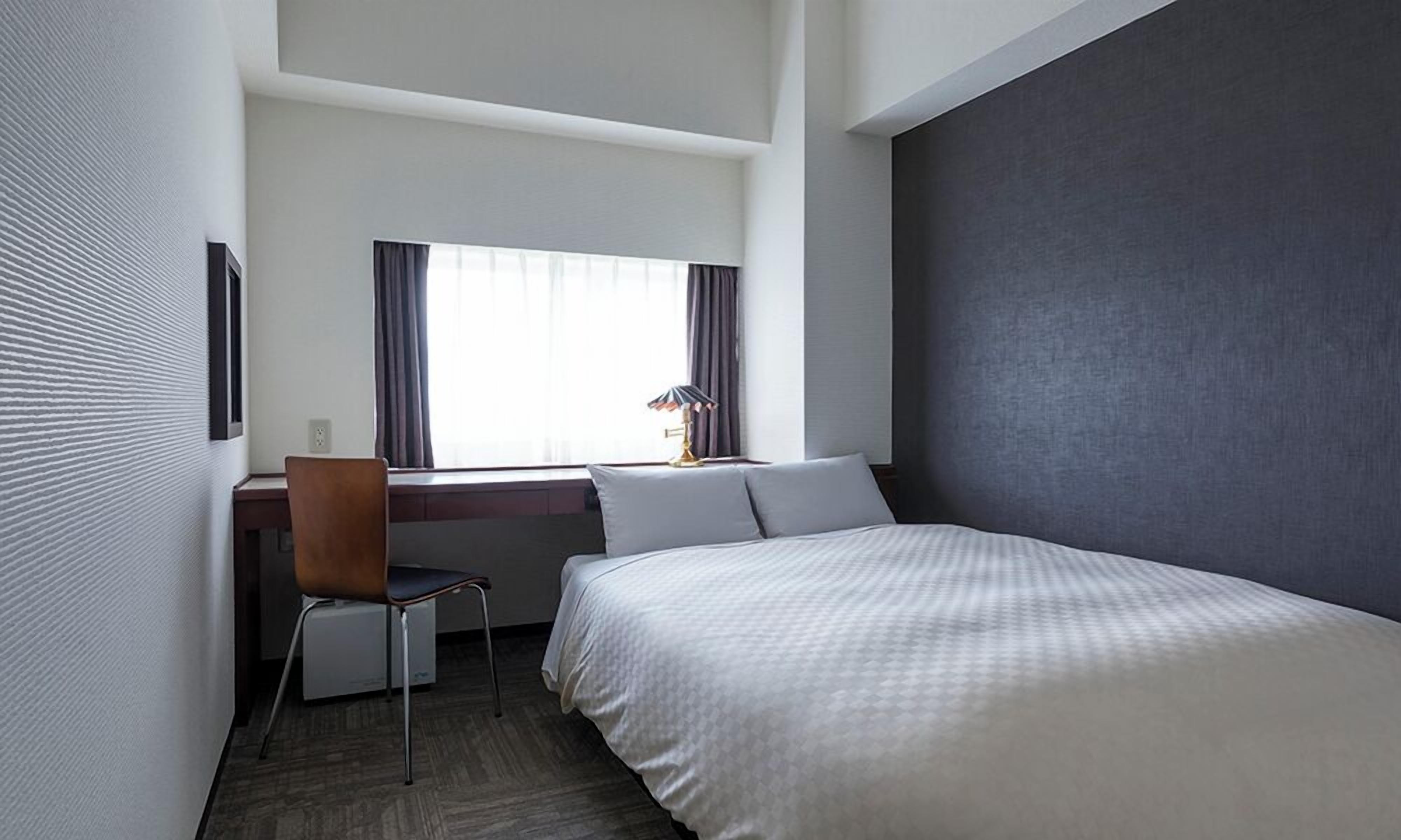 A hotel room with a bed, a desk with a chair, a window with curtains, and a lamp.