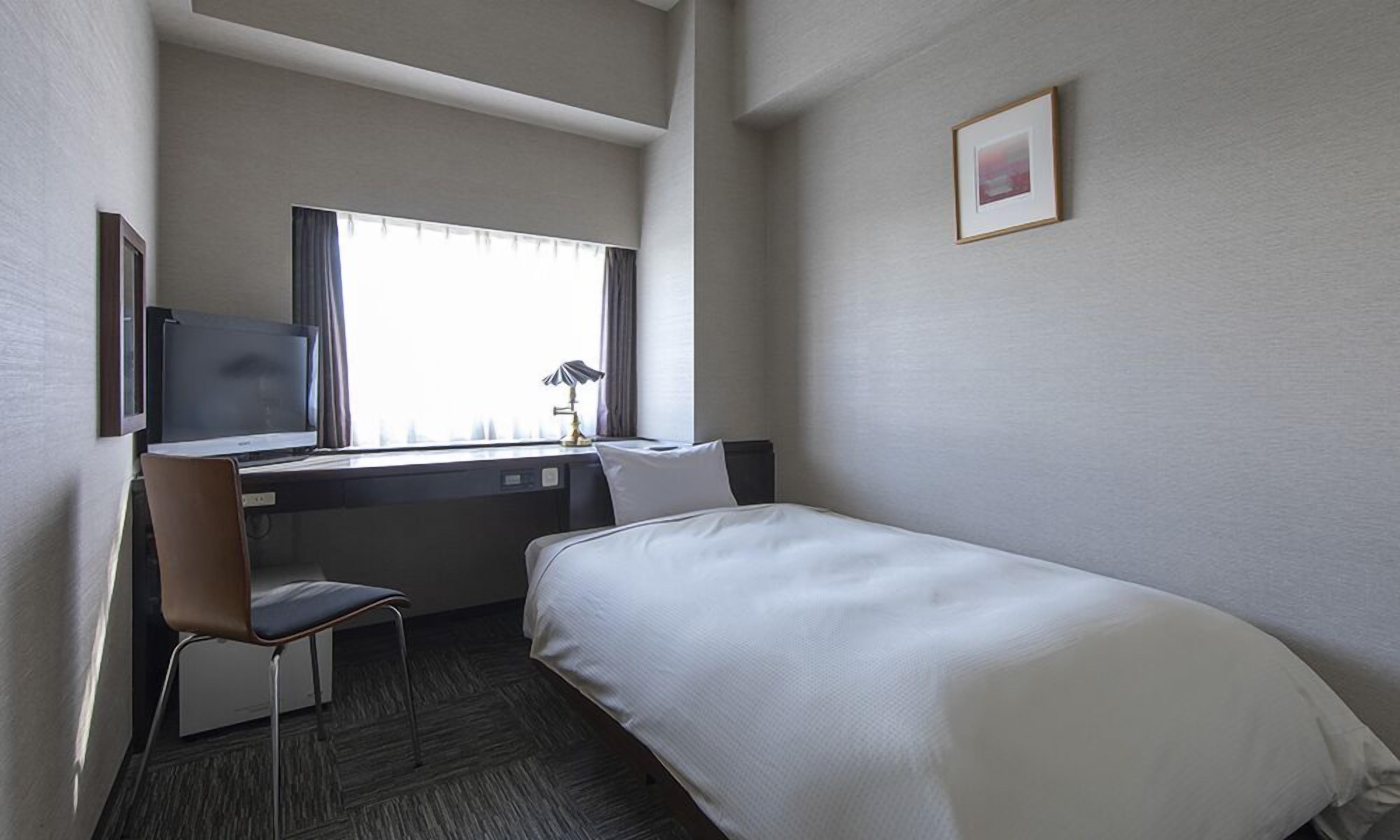 A hotel room with a bed, a desk, a chair, a TV, and a window with curtains.
