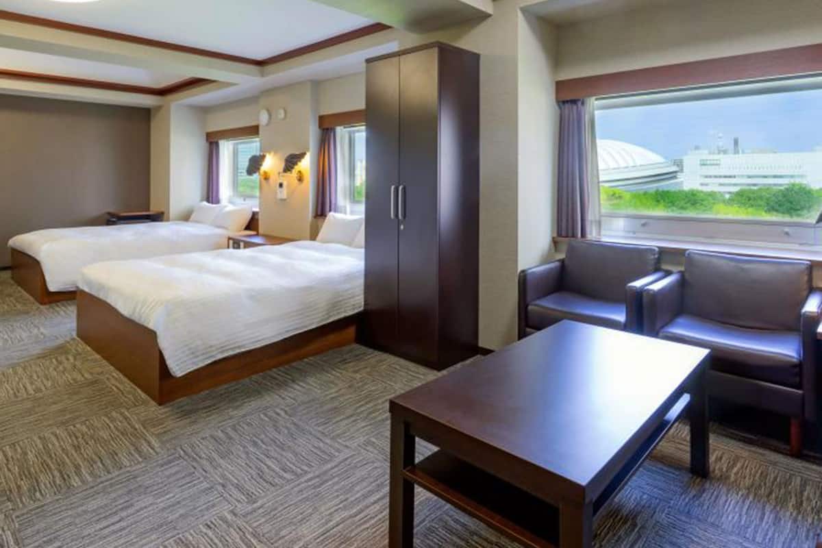 Une chambre d’hôtel avec deux lits, un bureau, un canapé et une grande fenêtre donnant sur un immeuble.