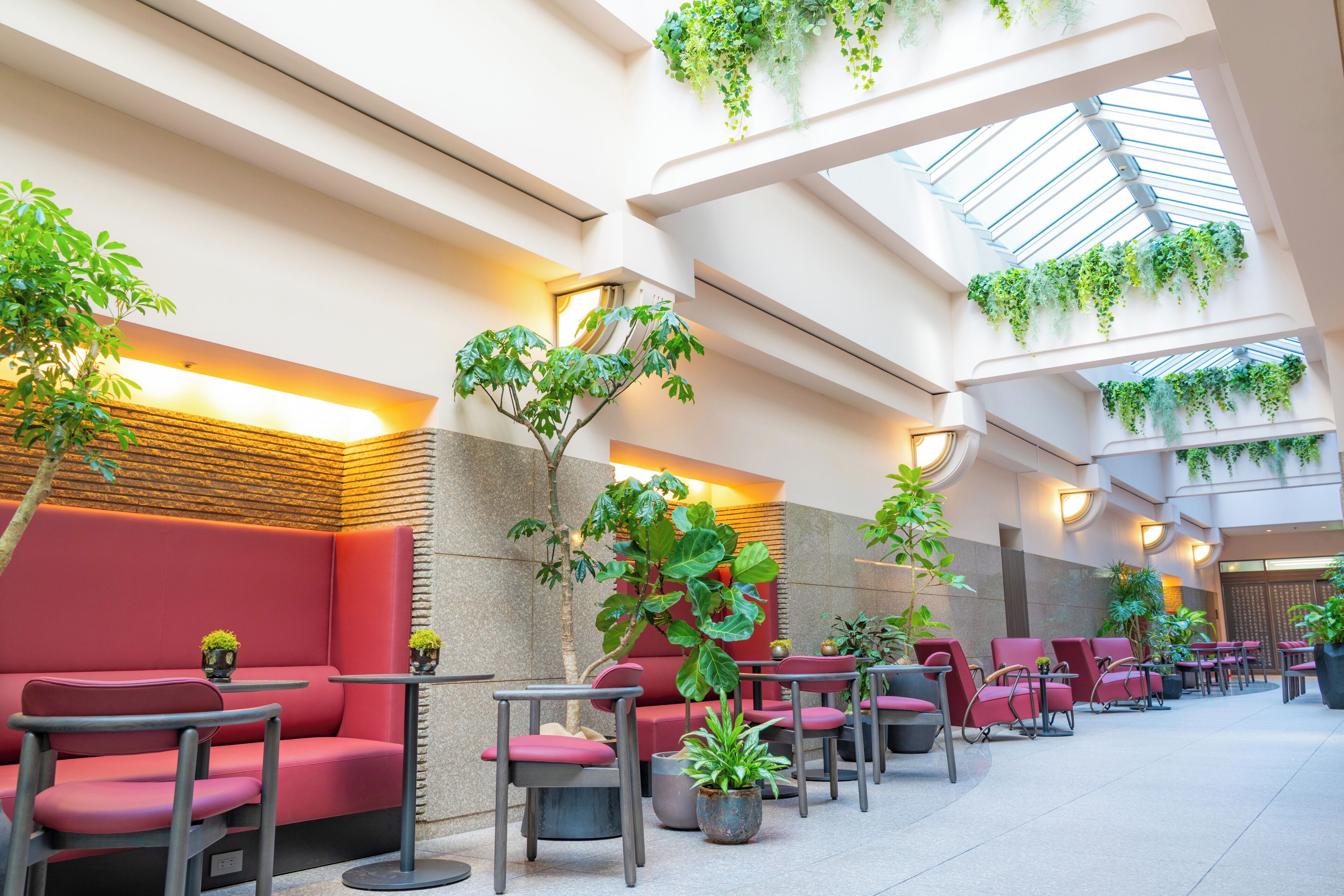 A modern indoor seating area with red cushioned benches and tables, surrounded by potted plants and a skylight.