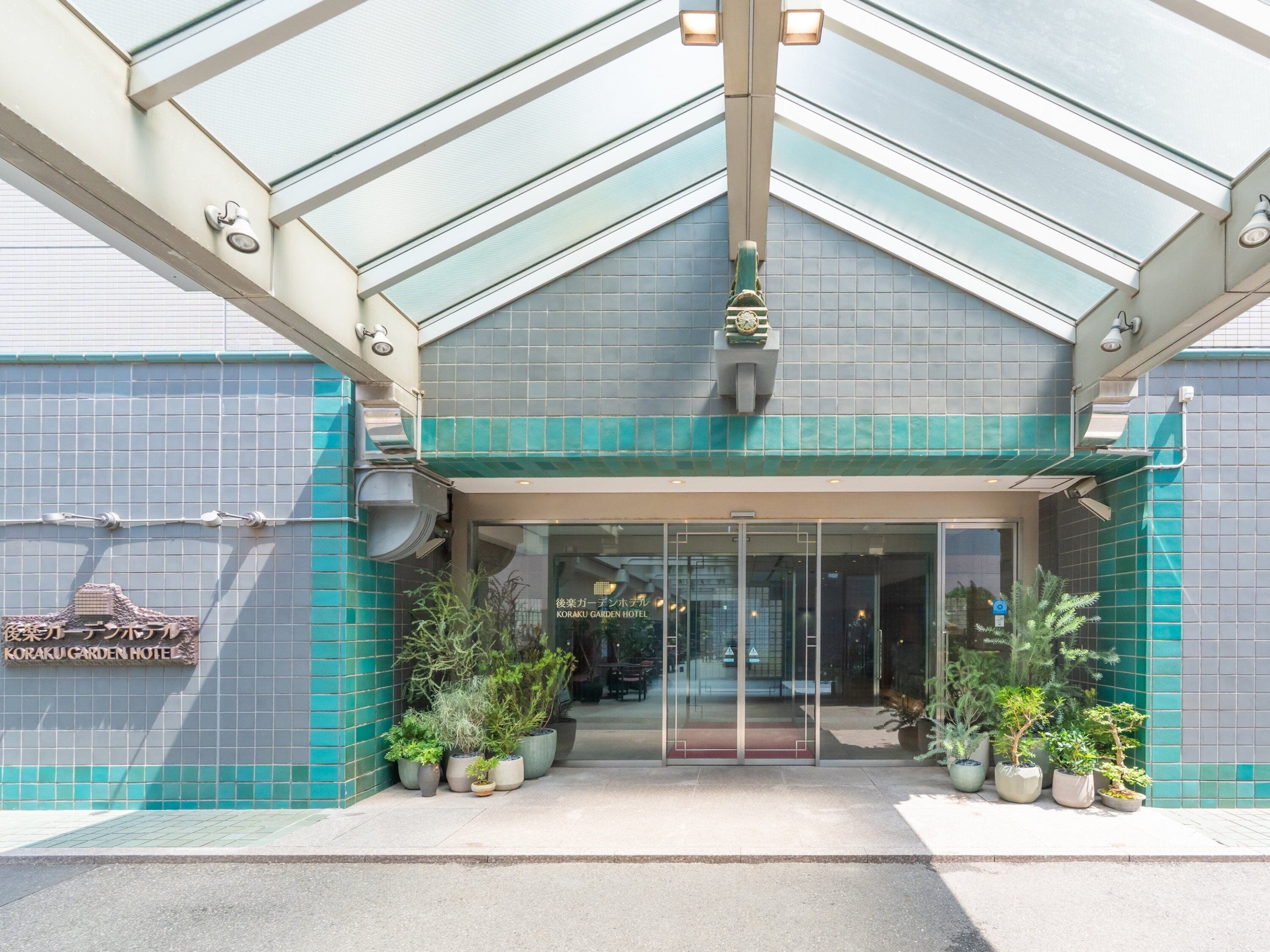 Entrada de hotel con puerta de vidrio, plantas en macetas y fachada con baldosas.