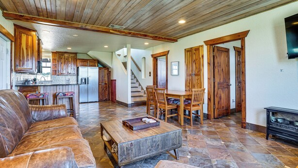 Amplia sala de estar con un sofá de cuero, una mesa de centro de madera y una mesa de comedor con sillas. Se observa una cocina al fondo con armarios de madera y un refrigerador.