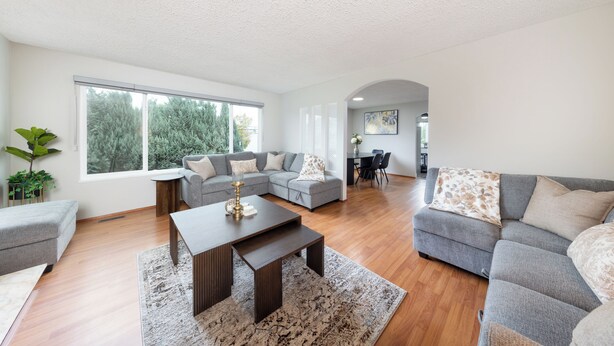 A spacious living room with a grey sectional sofa, a coffee table, and a large window offering a view of trees. - 3BR/2BA Home – 30 Mins to YVR, Family-Friendly (Delta)