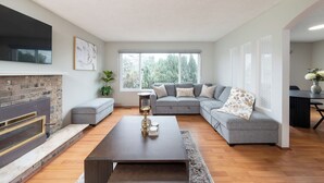 A modern living room with a fireplace, grey sofa, and a flat-screen TV mounted on the wall. - 3BR/2BA Home – 30 Mins to YVR, Family-Friendly (Delta)