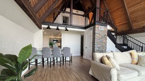 Amplia sala de estar con techo alto, vigas de madera, una escalera, una cocina al fondo y una gran planta en maceta.