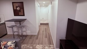 A modern living room with a flat-screen TV, a dining area with a table and two bar stools, and a hallway leading to a bedroom.
