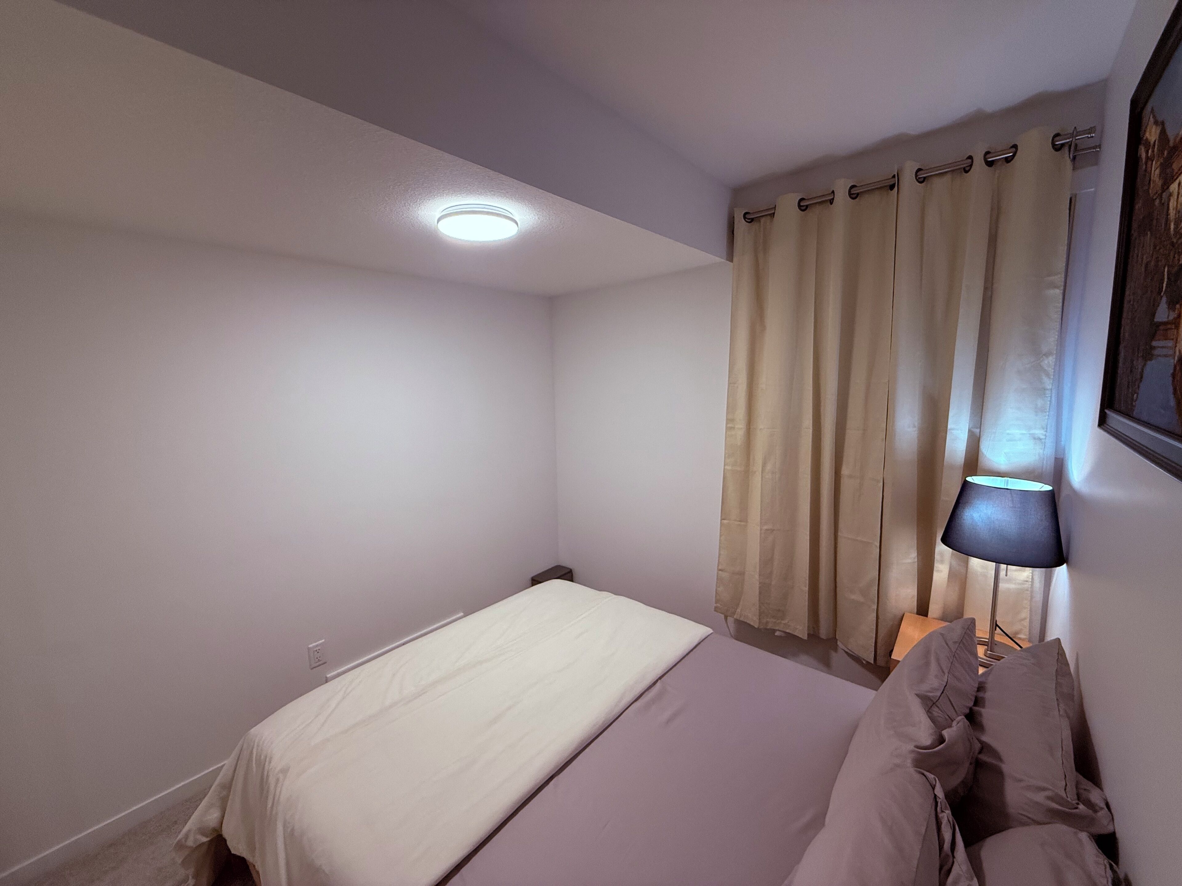 A small, neatly arranged bedroom with a single bed, a bedside lamp, and a window with curtains.