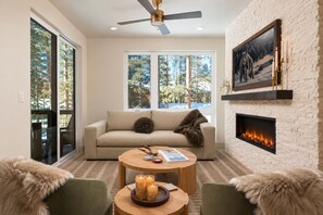 A modern living room with a fireplace, a sofa, a coffee table, and a large window offering a view of trees. - Luxe 3Br Townhome w/ Private Hot Tub, Sauna, Walk to Village and Gondola! (Dillon)