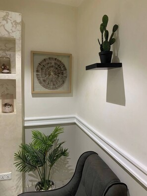 A corner of a room with a framed circular artwork, a potted cactus on a black shelf, a grey sofa, and a plant in a pot. - El Exclusive Apartment (Accra, East Legon.)