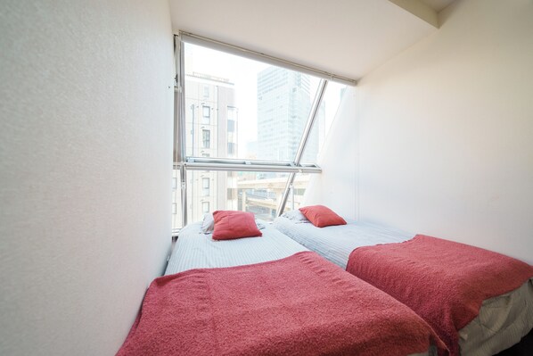 A small, bright room with two beds, each with a red blanket and pillow, and a large window offering a view of city buildings. - 6 min to Roppongi St! Spacious 5BR apt, 12 beds,140㎡,sleeps up to 12 guests. (Roppongi, Minato)
