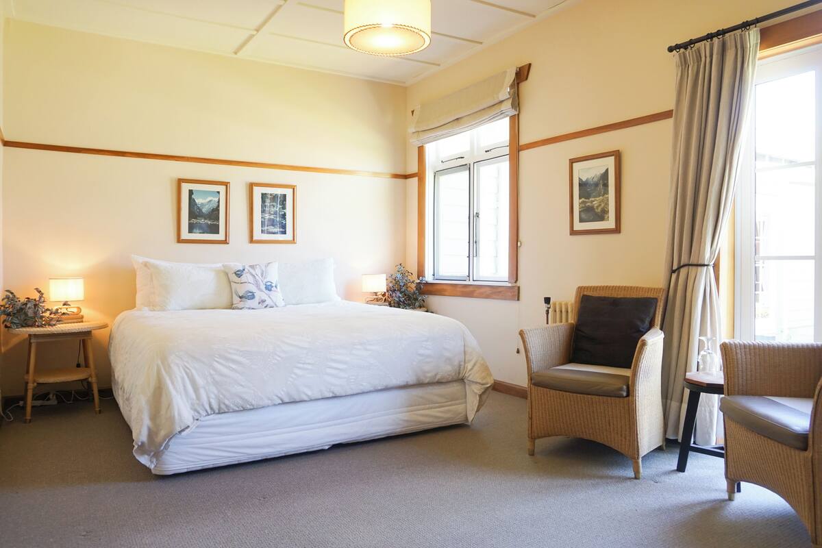 A hotel room with a large bed, two bedside tables, a chair, a window with curtains, and framed pictures on the wall.