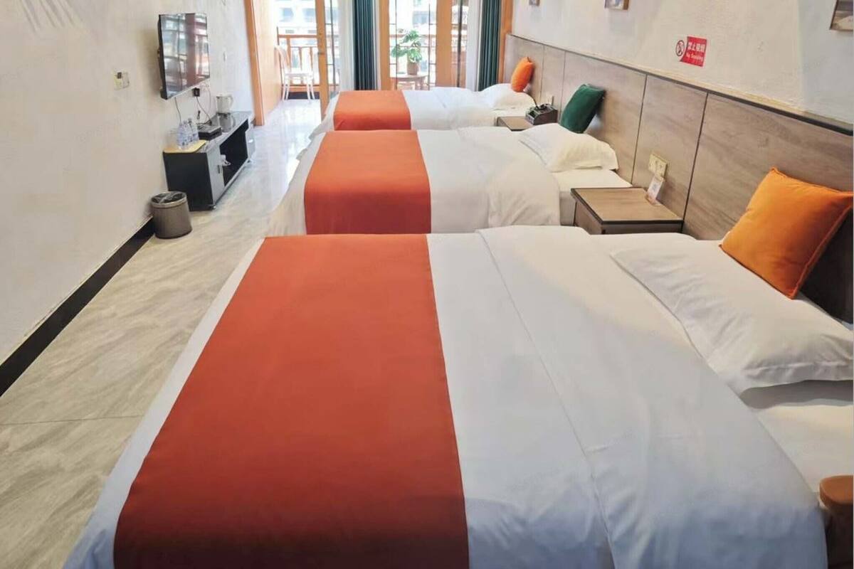 A hotel room with three beds, each with a white and orange bedspread. There is a small bedside table with a lamp, a TV mounted on the wall, and a trash can near the entrance.