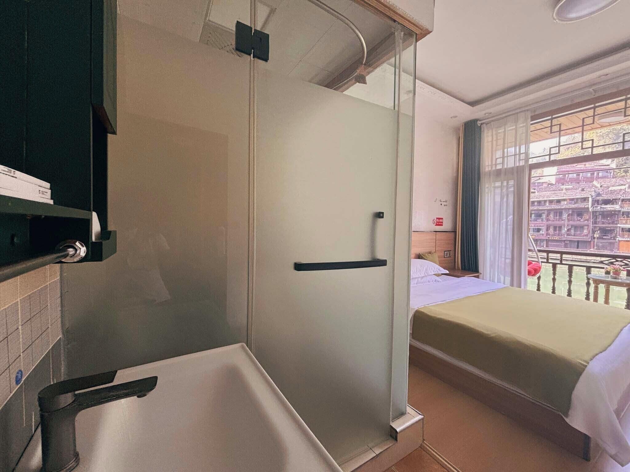 Habitación con ducha de vidrio, cama con cubrecama blanco y vistas a edificios tradicionales a través de una ventana.