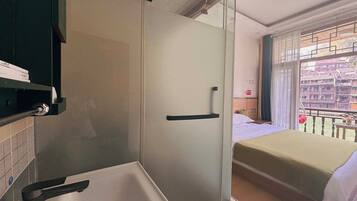 Habitación con ducha de vidrio, cama con cubrecama blanco y vistas a edificios tradicionales a través de una ventana.