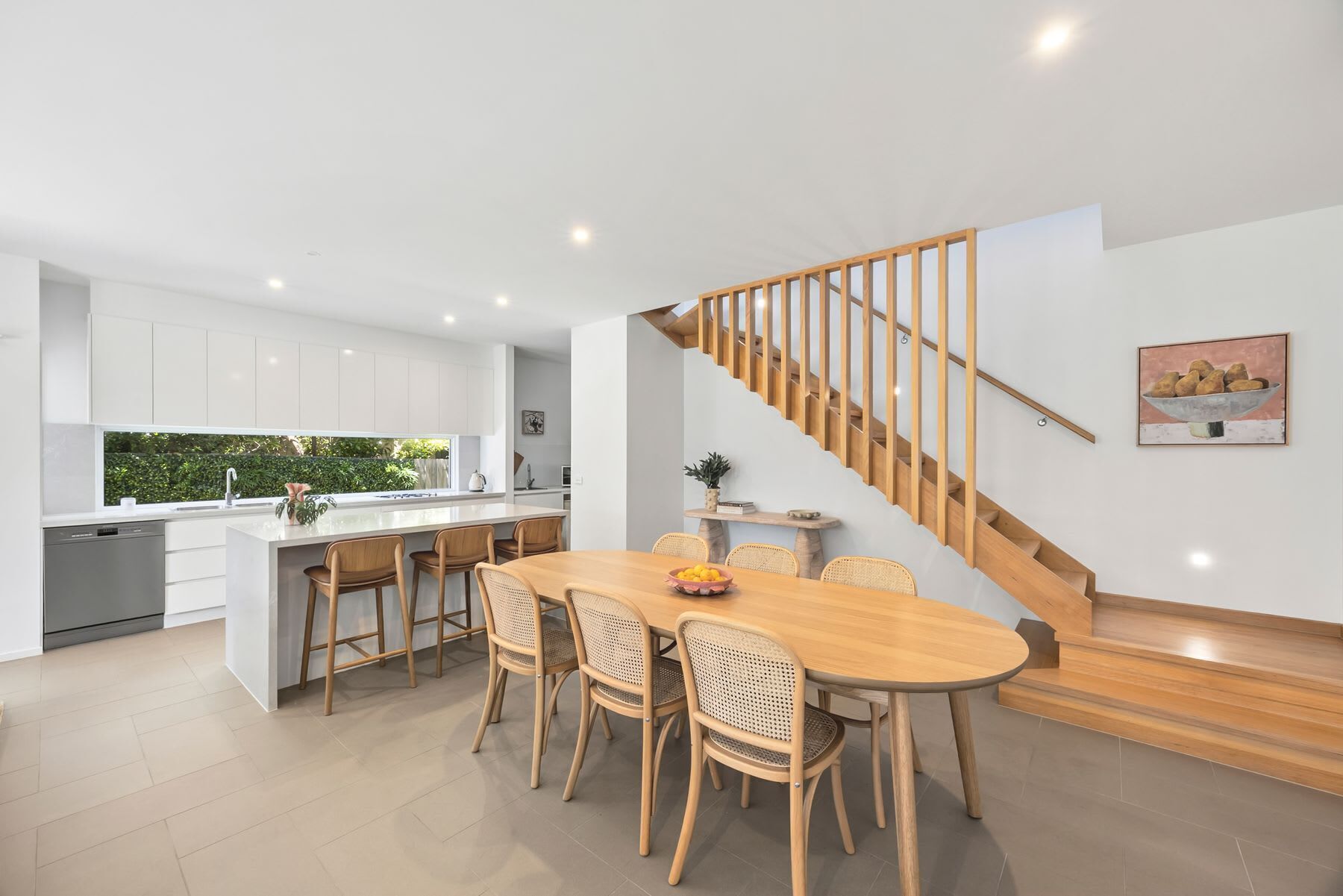 A modern kitchen with white cabinets, a wooden dining table with four chairs, and a staircase with wooden railings.