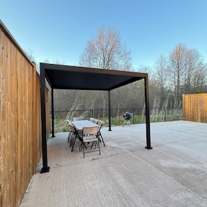 A covered patio area with a table and chairs, a grill, and a wooden fence.
