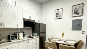 Een moderne keuken met witte kasten, een zwarte magnetron en een koelkast. Er staat een eettafel met twee stoelen bij, en een vaas met gedroogde planten.