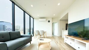 Un salon moderne avec un canapé, une table basse ronde, un téléviseur posé sur un meuble en bois, et une vue sur la ville grâce à de grandes fenêtres.