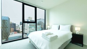 Une chambre d’hôtel moderne dotée d’un grand lit, d’une table de chevet, d’une lampe et offrant une vue sur la ville grâce à de grandes fenêtres du sol au plafond.