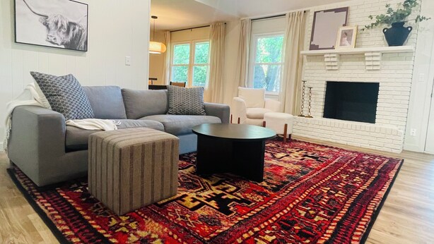 Ett modernt vardagsrum med en grå soffa, en randig pall, ett runt soffbord och en röd, mönstrad matta. Det finns en vit öppen spis med en svart insats, en inramad tavla ovanför och en krukväxt på spiselkransen.