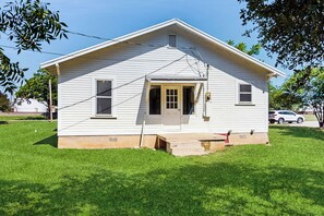 A single-story white house with a front door, a small porch, and a grassy yard. - Walkable 1900s Home w/ Porch in Downtown Buda (Buda)