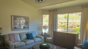 A living room with a beige sofa, a coffee table with a green centerpiece, a patterned rug, a painting on the wall, and a view of a garden through a large window. - Mountain-View Backyard Oasis! King City Getaway (King City)