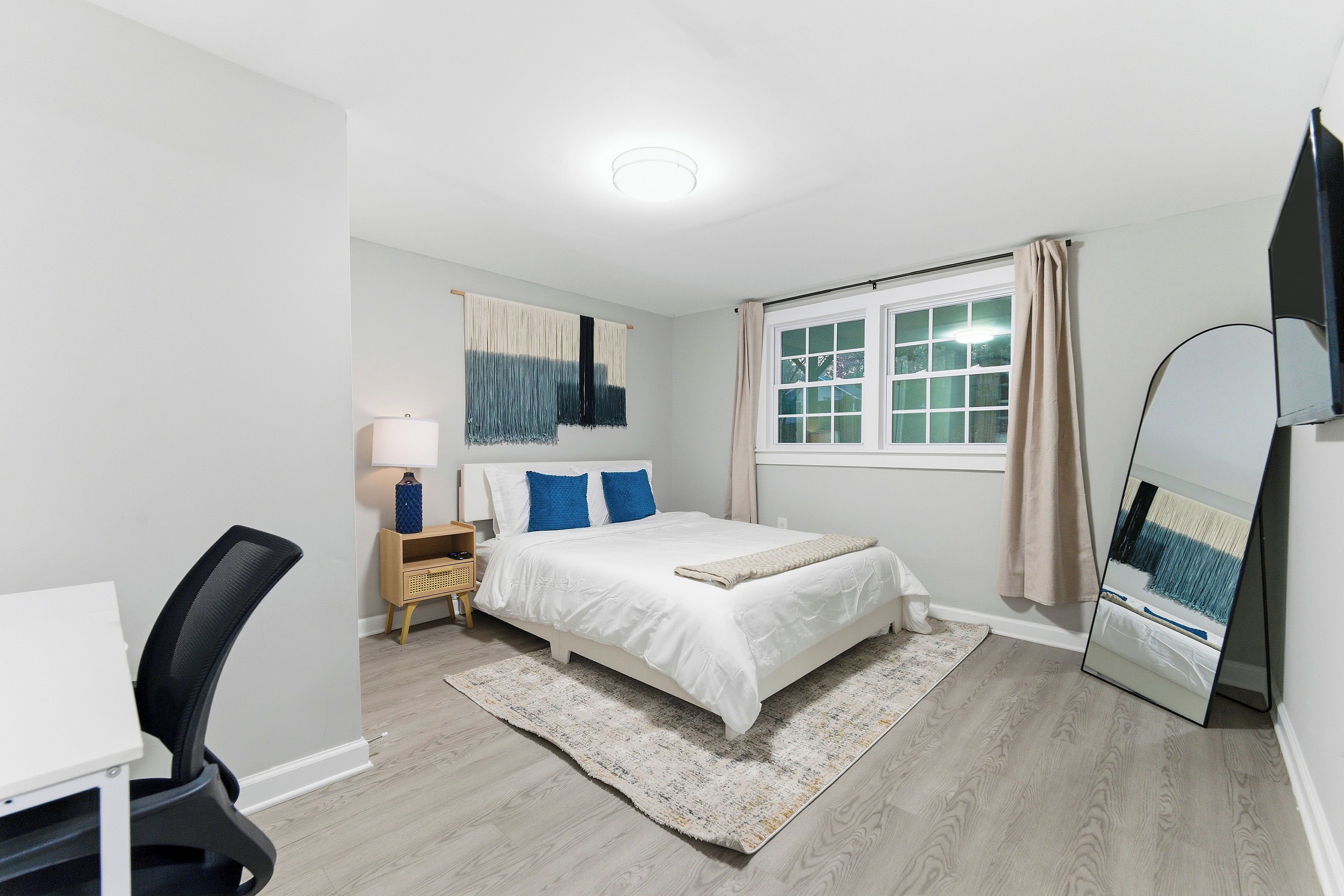 Une chambre à coucher comprenant un lit, un bureau, une chaise, une lampe, un miroir et une fenêtre avec des rideaux.