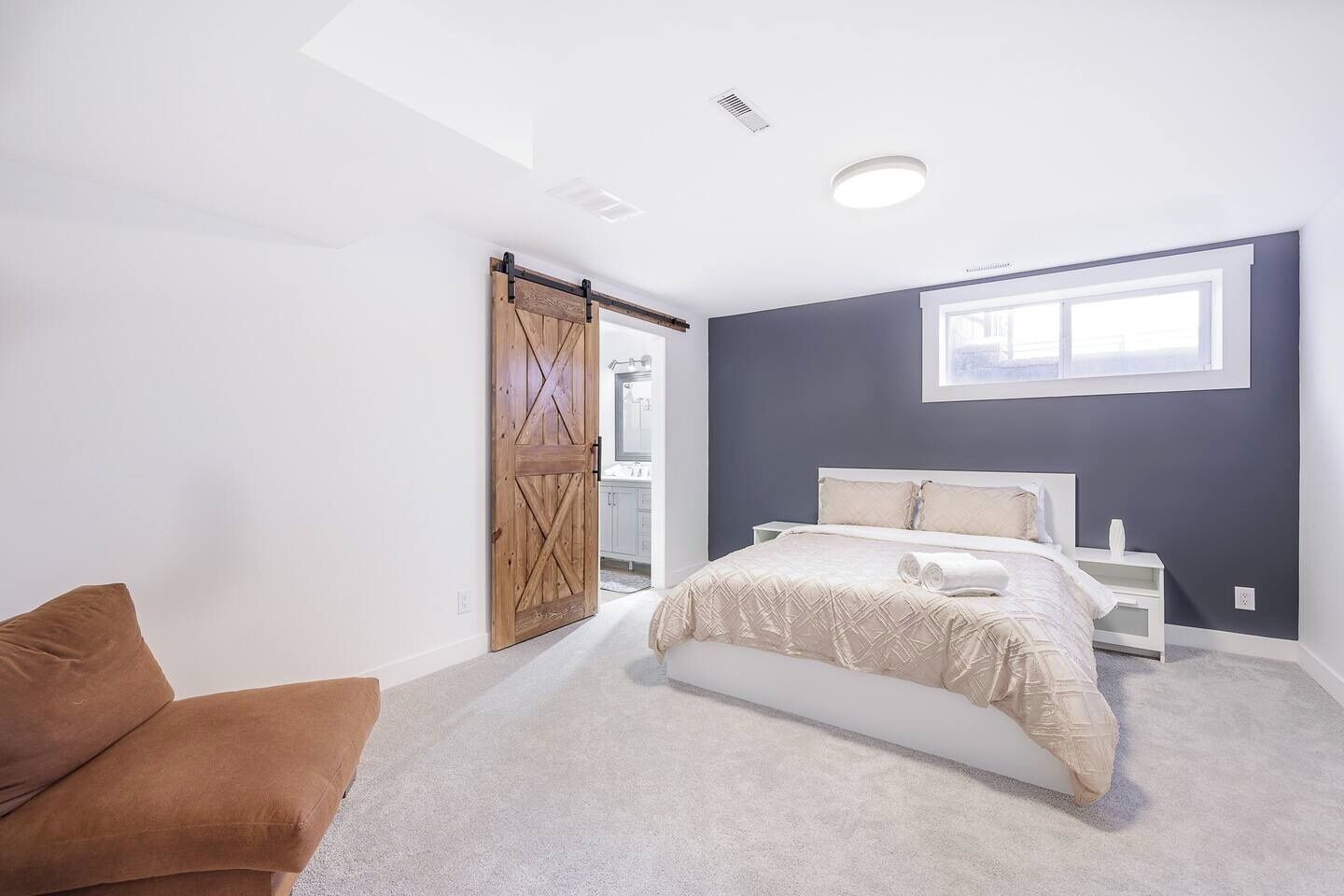 Een slaapkamer met een bed, een bruine stoel, een groot raam en een deur naar een badkamer.