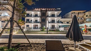 Un bâtiment moderne à plusieurs étages, doté de balcons, une zone sablonneuse avec un fauteuil de relaxation et un parasol, et un arbre au premier plan.