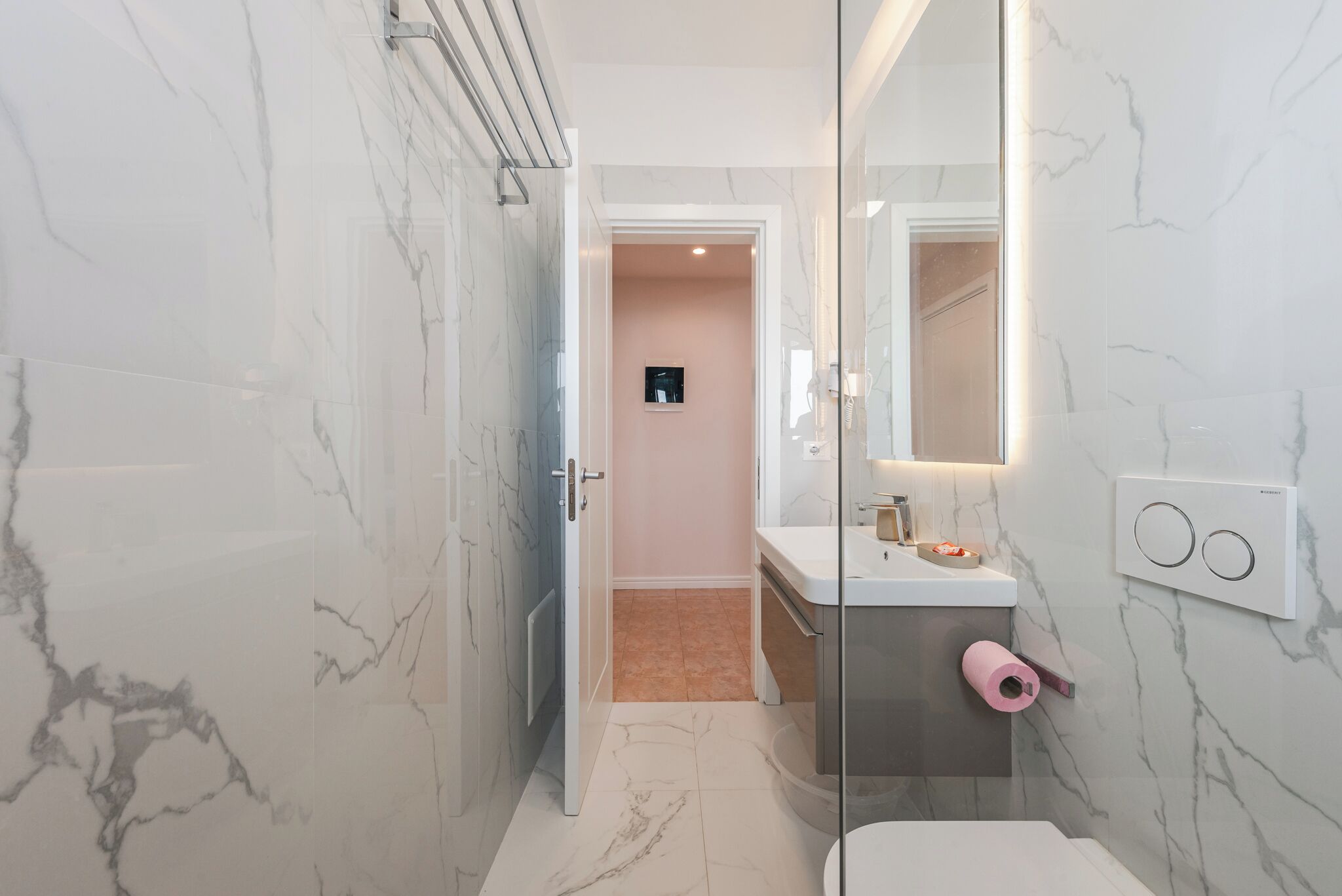 Un baño moderno con paredes de mármol, cabina de ducha con mampara de vidrio, lavamanos blanco y retrete.