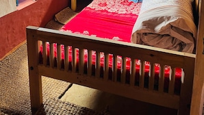 Een houten bed met een bloemenbedekking in rood en wit, een klein nachtkastje en een raam met blauwe luiken.