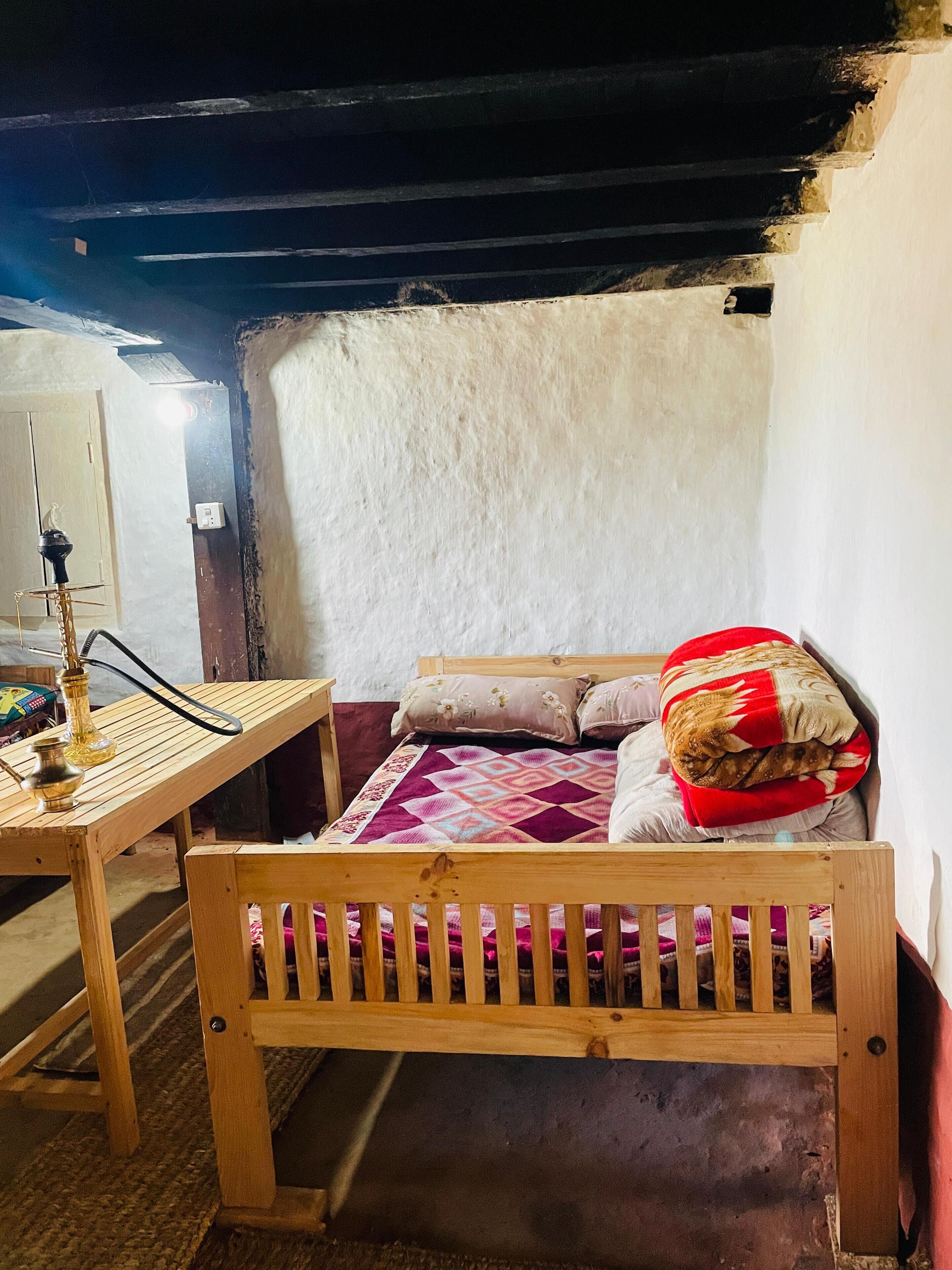 A small, rustic room with a wooden bed, a table with a hookah, and a wall with a rough texture.