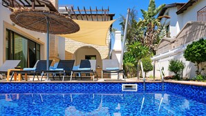 Une piscine aux carreaux de mosaïque bleus, des chaises longues et une pergola surmontée d’un parasol en chaume.