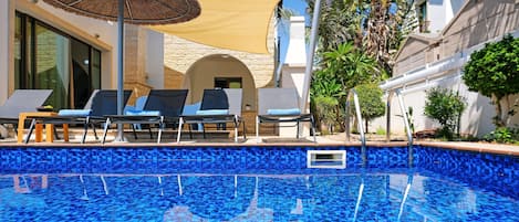 En swimmingpool med blå mosaikplattor, solstolar och en pergola med ett halmtäckt parasoll.