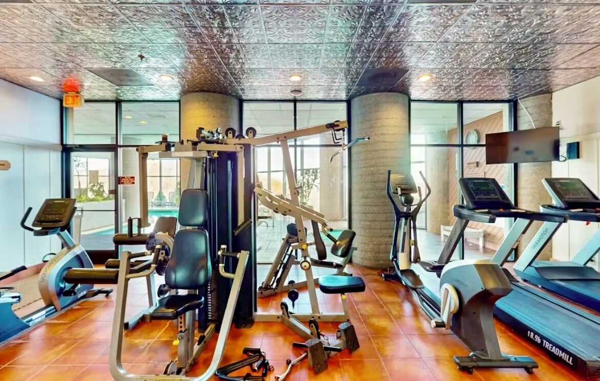 A gym with various exercise equipment, including treadmills and weight machines, and large windows allowing natural light.