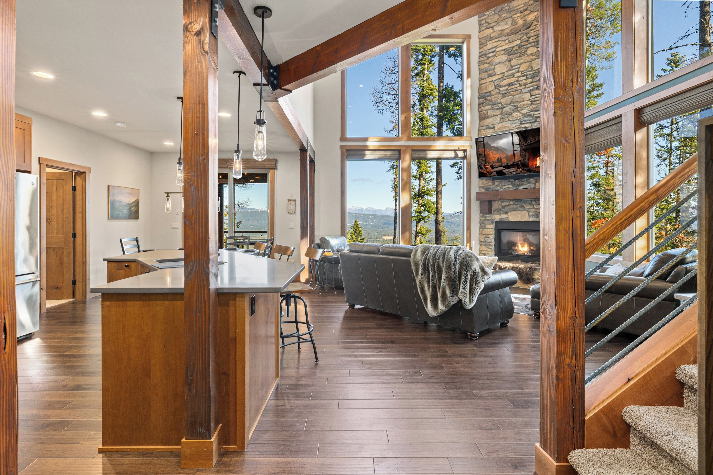 A spacious living area with a stone fireplace, a large window overlooking mountains, and a wooden staircase.