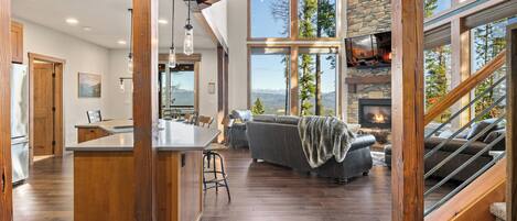 A spacious living area with a stone fireplace, a large window overlooking mountains, and a wooden staircase.