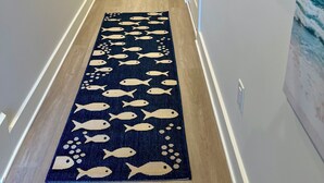 En hall med ett blått mattgolv som har ett fiskmönster, vita väggar och en dörr som leder till ett rum med ett fönster och en målning av segelbåtar.