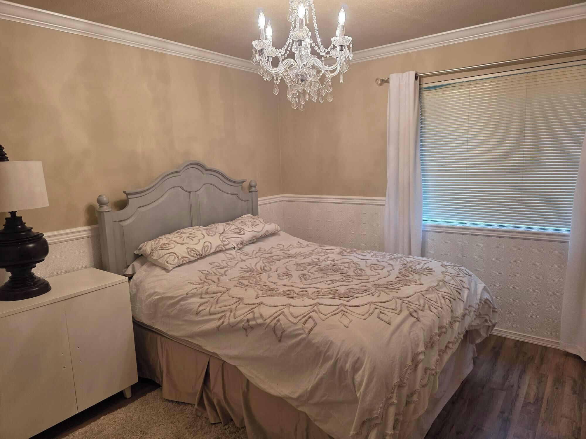 A bedroom with a bed, a nightstand, a lamp, a chandelier, and a window with blinds.