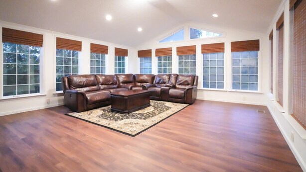 A spacious living room with a large L-shaped leather sectional sofa, a matching ottoman, and a patterned rug. The room has large windows with blinds, wooden flooring, and recessed ceiling lighting.