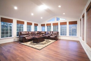 A spacious living room with a large L-shaped leather sectional sofa, a matching ottoman, and a patterned rug. The room has large windows with blinds, wooden flooring, and recessed ceiling lighting. - Farm Stay at Golden Meadows (Concord)