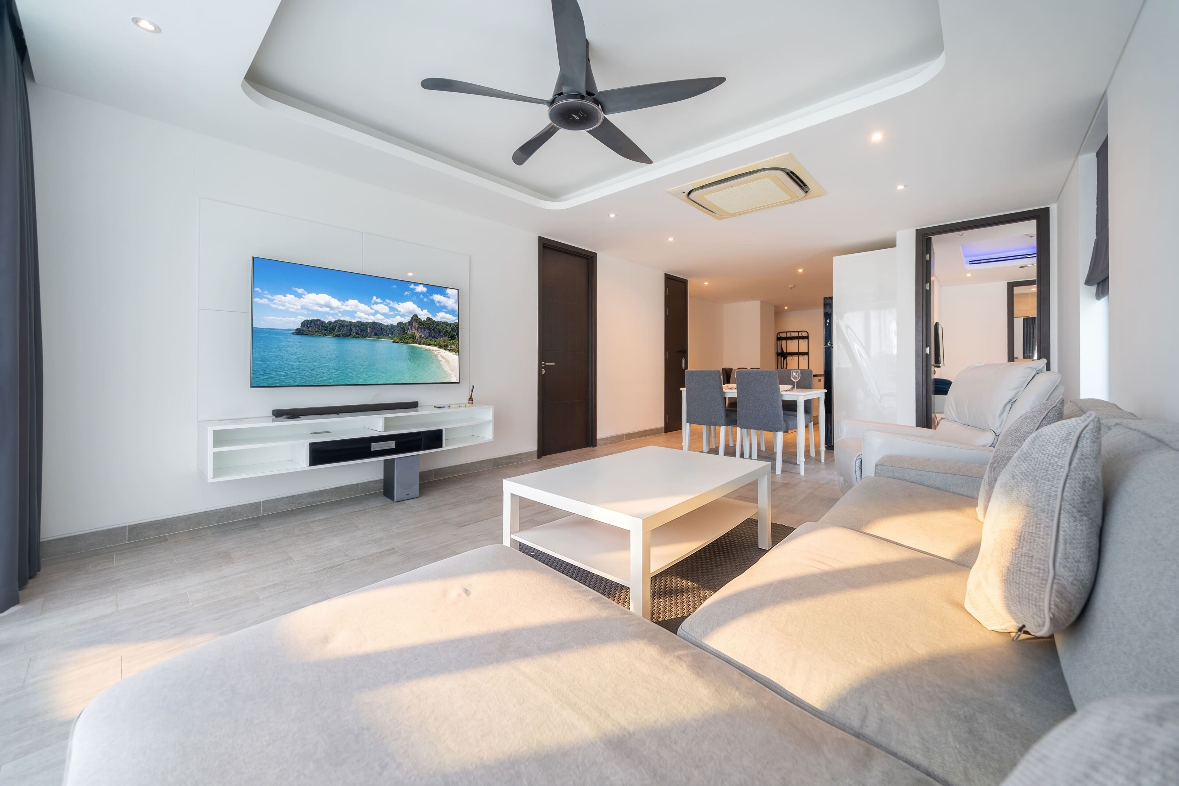 Una sala de estar moderna con un televisor de pantalla plana, un ventilador de techo, un sofá blanco y un comedor con sillas.