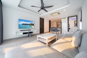 Egy modern nappali, melyben található egy síkképernyős televízió, egy mennyezeti ventilátor, egy fehér kanapé és egy étkezősarok székekkel.