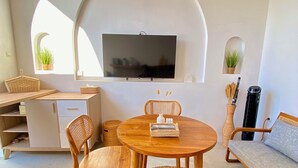 Ein modernes Wohnzimmer mit einem runden Holztisch, zwei Korbstühlen, einer Couch, einem an einer weißen Wand montierten Fernseher und einem kleinen Topfpflanzen.
