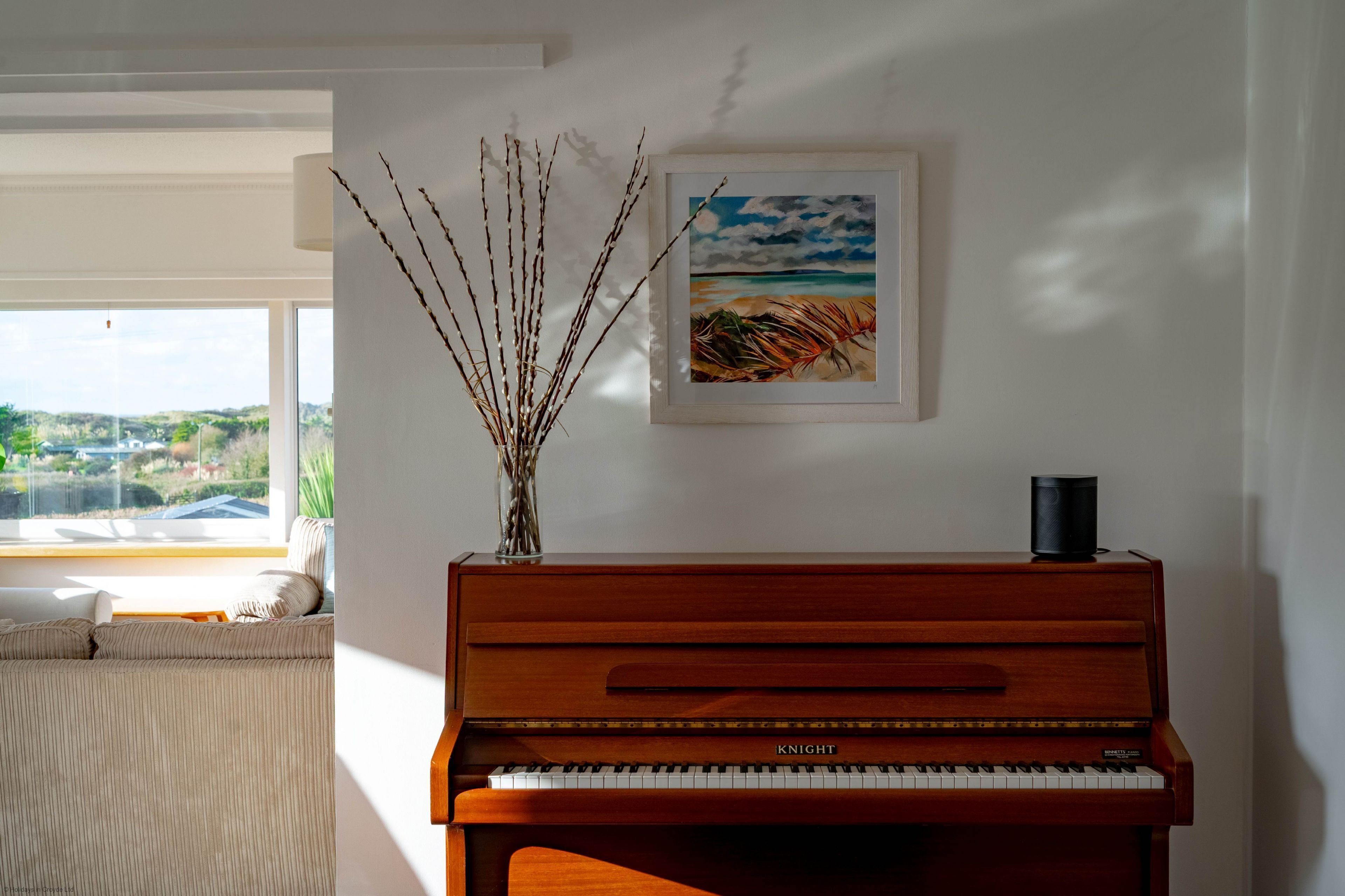 A piano with a framed painting above it, a vase with branches, and a view of a landscape through a window.